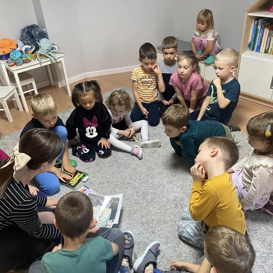 alt="Zajęcia edukacyjne w przedszkolu SunnyKids w Krakowie - dzieci w wieku 3-6 lat zgromadzone w kółku z nauczycielką podczas czytania książki, rozwijanie umiejętności językowych w placówkach na Żabińcu i Górce Narodowej